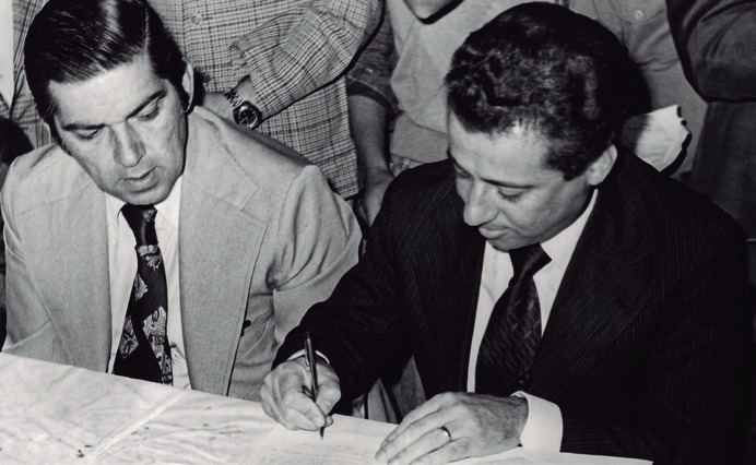 Dois homens &agrave; mesa, um deles assinando um documento