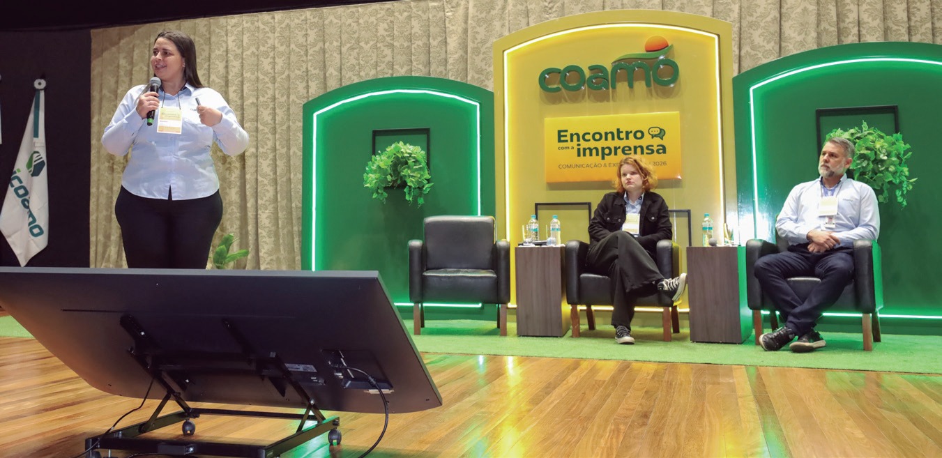 Mulher em p&eacute; no palco falando ao p&uacute;blico, com dois participantes sentados ao fundo