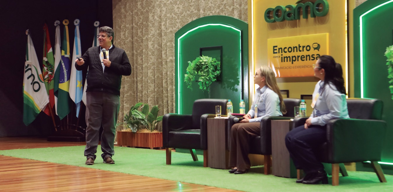 Homem em p&eacute; no palco apresentando, ao lado de duas pessoas sentadas