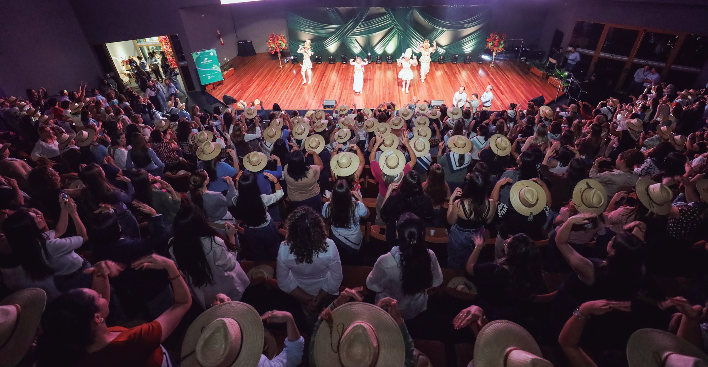 Plateia de mulheres de chap&eacute;u diante do palco com apresenta&ccedil;&atilde;o art&iacute;stica