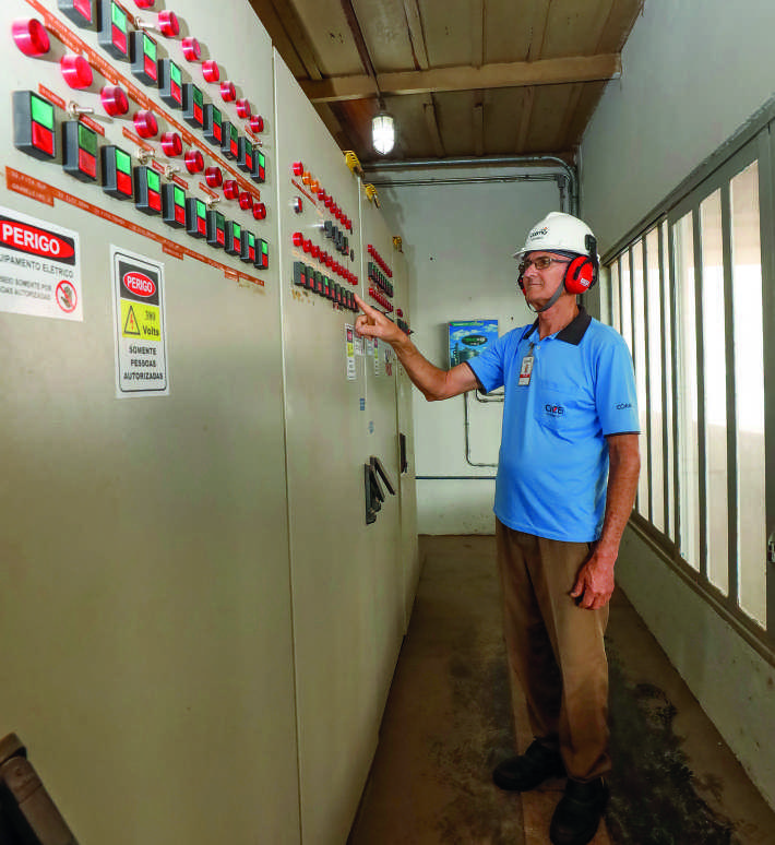 Funcionário da Coamo, de capacete e protetor auricular, opera um grande painel elétrico industrial.